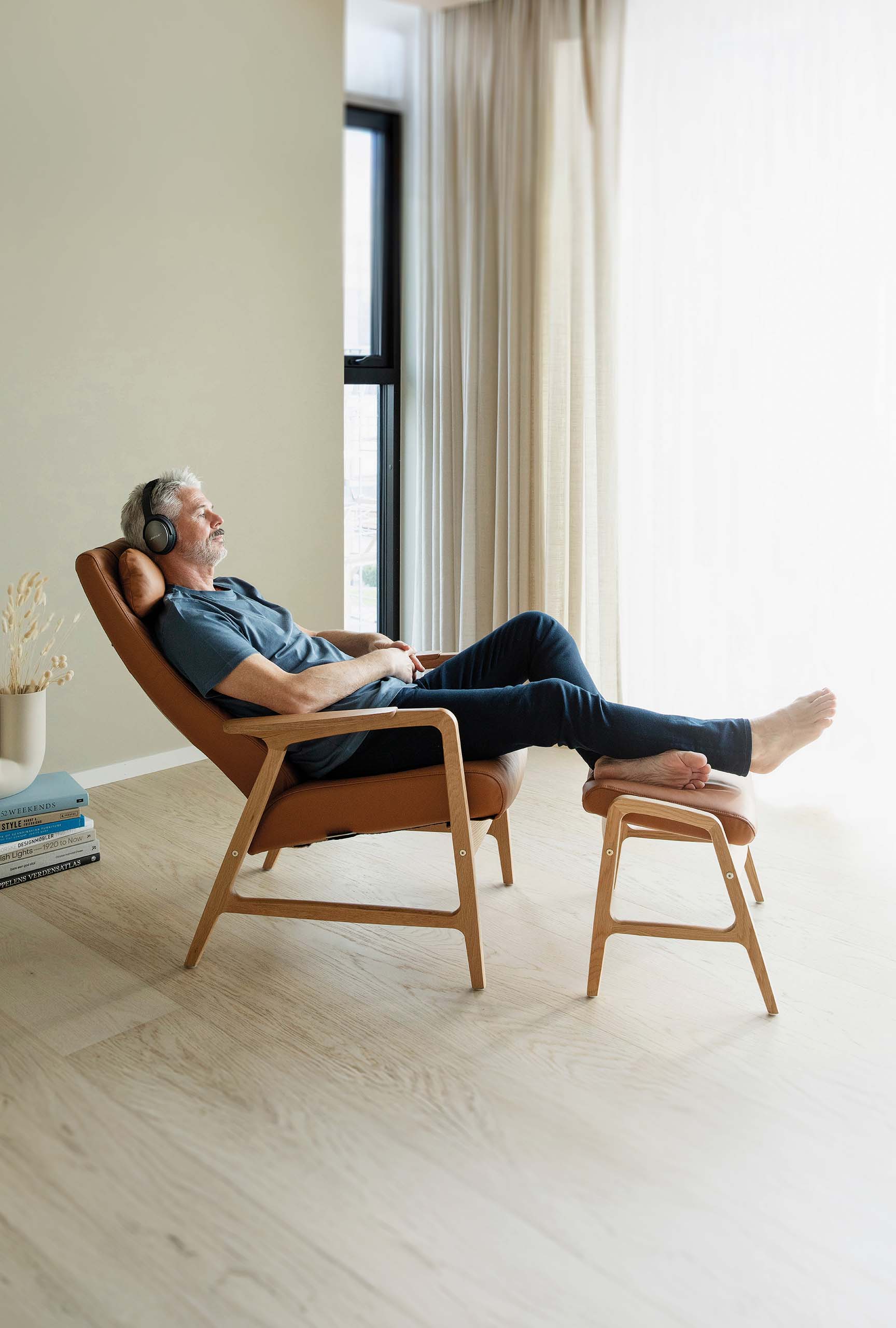 „Person entspannt in einem Lounge‑Sessel mit Fußhocker aus Holz und cognacfarbener Polsterung in einem hellen, minimalistischen Raum mit großem Fenster.