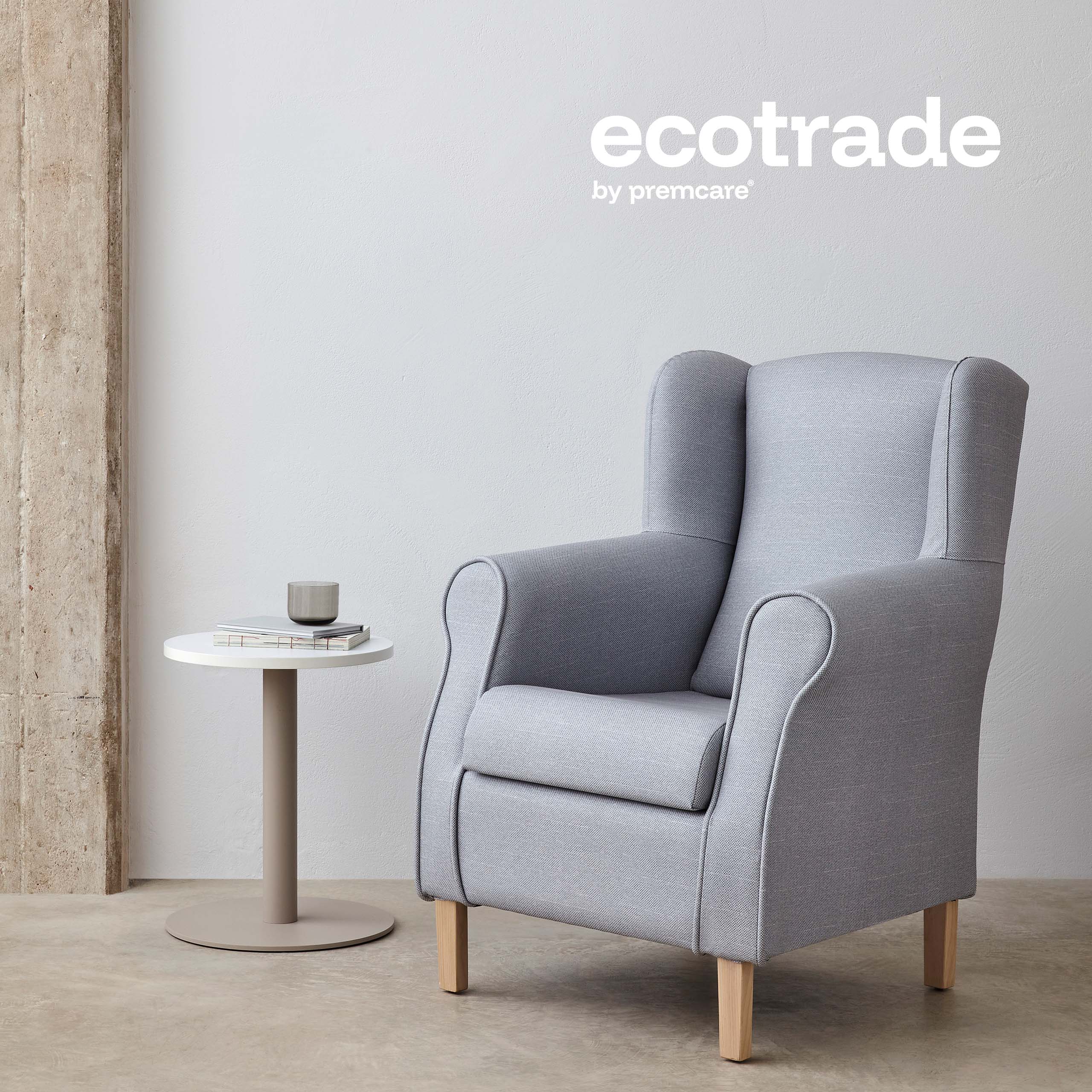 Hellblau gepolsterter Ohrensessel mit Holzfüßen neben einem runden Beistelltisch mit zwei Büchern und einem Trinkglas in einem modernen Raum mit Betonoptik; im Bild ist das Logo ‚ecotrade by premcare‘ zu sehen.