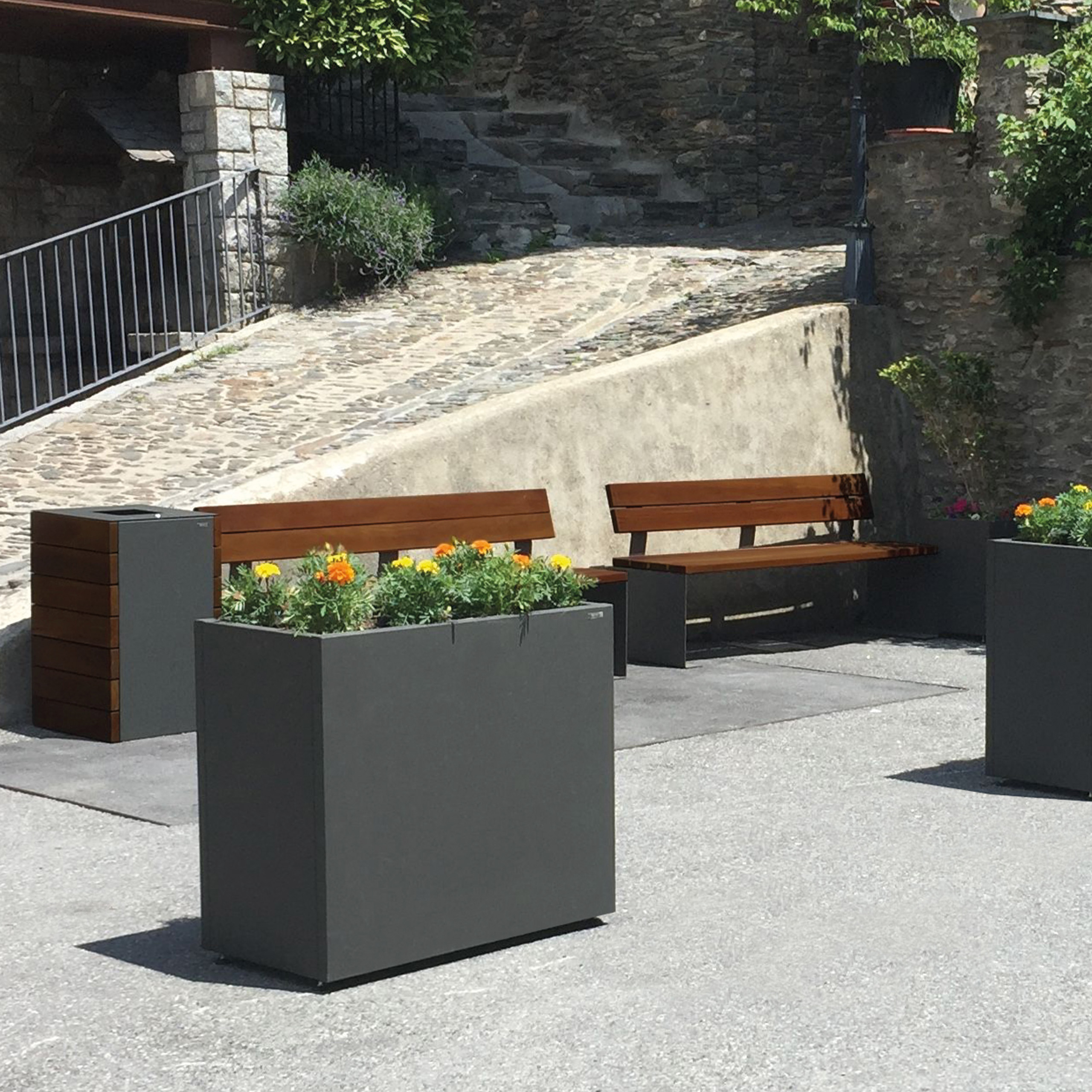 Zwei hölzerne Sitzbänke und ein passender Mülleimer auf einer städtischen Fläche, ergänzt durch zwei Blumenkübel mit gelben und orangefarbenen Pflanzen.
