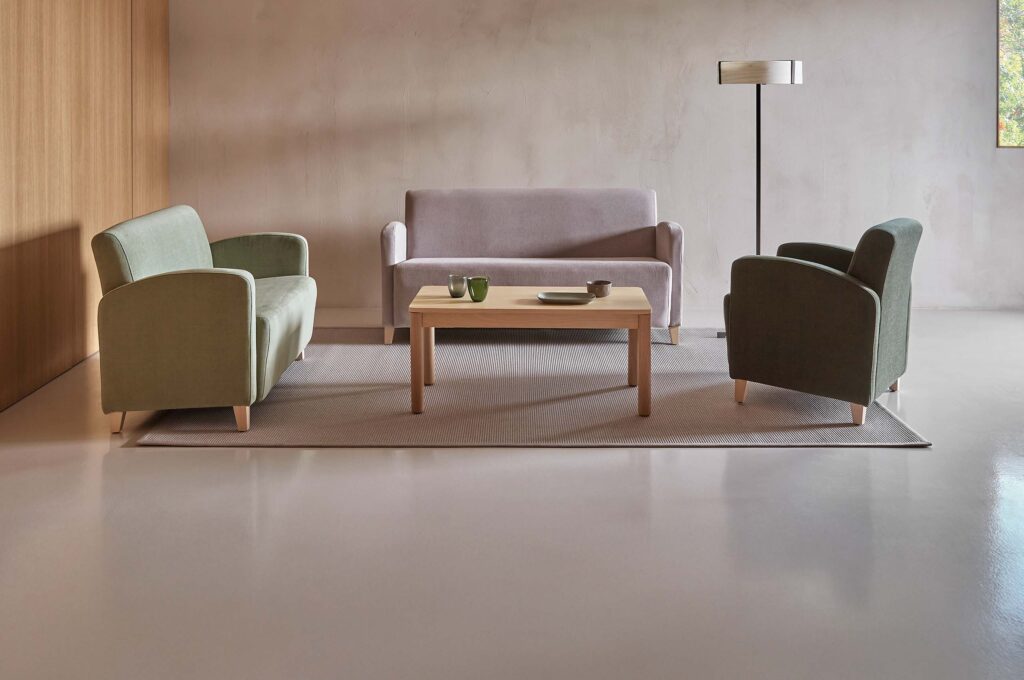 Moderner Wohnbereich mit hellem Holztisch, drei farblich abgestimmten Sitzmöbeln und einer schlichten Stehleuchte vor beige‑holzigem Hintergrund.