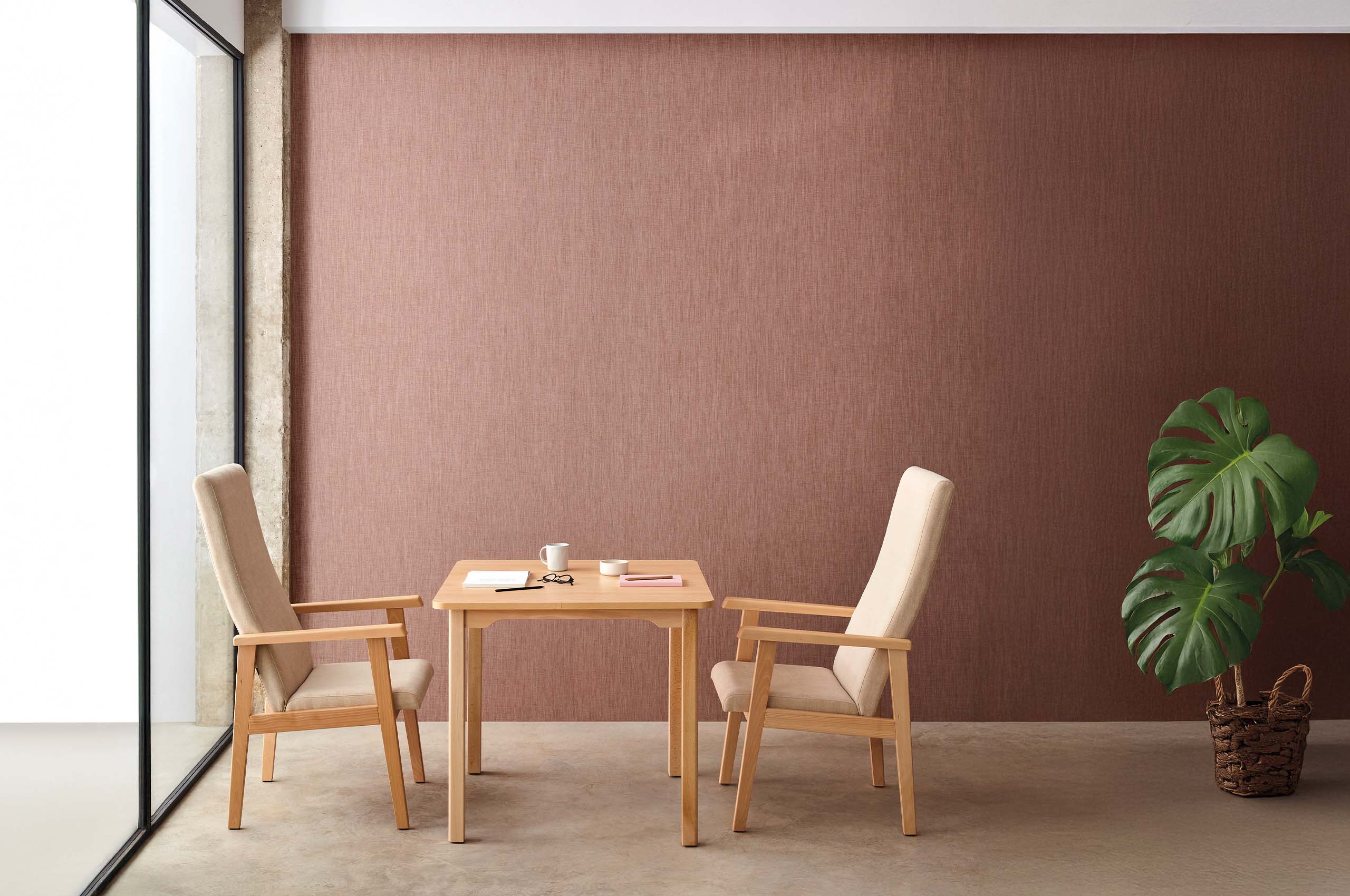 Minimalistischer Innenraum mit hellem Holztisch, zwei gepolsterten Holzstühlen und einer großen Monstera‑Pflanze vor warmem rotbraunem Hintergrund.