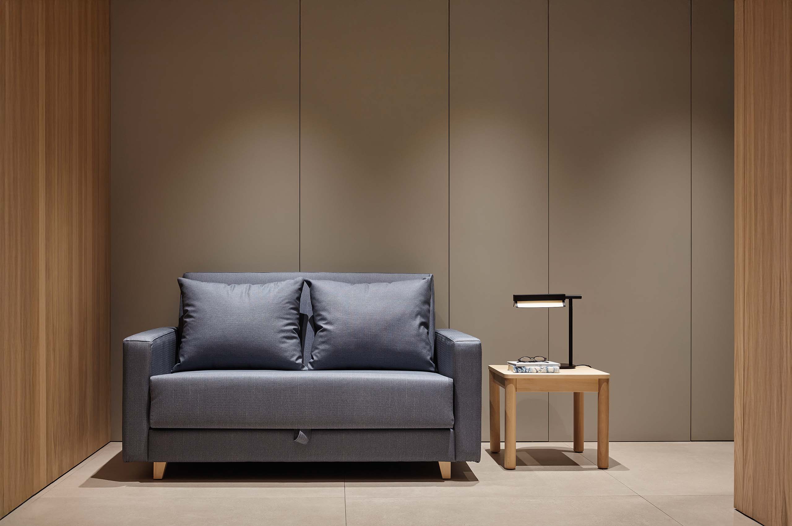 Modernes, minimalistisches Interieur mit grauem Sofa, hellem Beistelltisch, schwarzer Tischlampe sowie warmen Holzpaneelen und taupefarbener Wand.